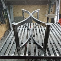 WANDZEL CUSTOM MICHAŁ WANDZEL - Dwie metalowe, surowe nogi do stołu o łukowatym kształcie, ustawione na metalowym stole warsztatowym z widocznymi śladami obróbki.