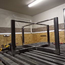 WANDZEL CUSTOM MICHAŁ WANDZEL - Surowa, metalowa konstrukcja w kształcie sześcianu, spawana na metalowym stole warsztatowym w jasnym oświetleniu, szlifierka kątowa leży obok.