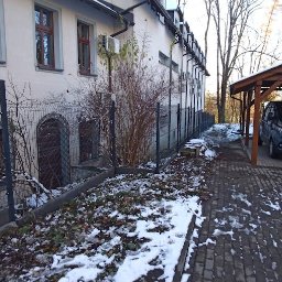 WANDZEL CUSTOM MICHAŁ WANDZEL - Szara siatka ogrodzeniowa z metalowymi słupkami, częściowo pokryta śniegiem, biegnąca wzdłuż budynku z licznymi klimatyzatorami na ścianie i wiatą garażową z zaparkowanym samochodem w tle.