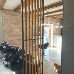 Metalowa ścianka działowa z drewnianymi listwami w salonie fryzjerskim, oddzielająca stanowiska mycia włosów od reszty przestrzeni, z widocznymi belkami stropowymi i ceglaną ścianą w tle.