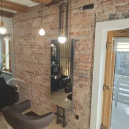 Wnętrze salonu fryzjerskiego z ceglaną ścianą, metalową konstrukcją lustra i drewnianymi belkami na suficie, oświetlone wiszącymi żarówkami.