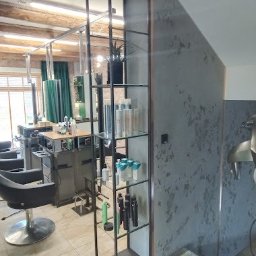 WANDZEL CUSTOM MICHAŁ WANDZEL - Metalowa regałowa konstrukcja z półkami szklanymi, wypełniona kosmetykami w salonie fryzjerskim, widoczny fragment stanowiska pracy i lustra.