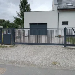 Nowoczesna, przesuwna brama wjazdowa w kolorze grafitowym, z pionowymi szczeblami, na tle domu z garażem i szarą nawierzchnią z kamienia.