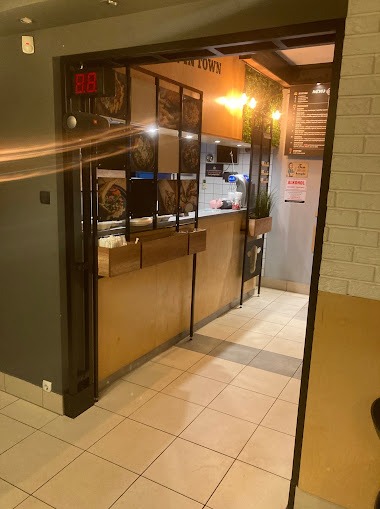 Wejście do lokalu gastronomicznego z metalową konstrukcją i drewnianym blatem, widoczne menu i dekoracje roślinne.