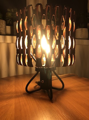 Ręcznie robiona lampka nocna z metalowym ażurowym kloszem i podstawą z czarnej liny, oświetlona ciepłym światłem żarówki, stoi na drewnianym blacie.