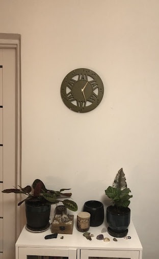 Dekoracyjny zegar ścienny w kolorze oliwkowym z rzymskimi cyframi, wiszący nad białą komodą z roślinami doniczkowymi i ozdobami.