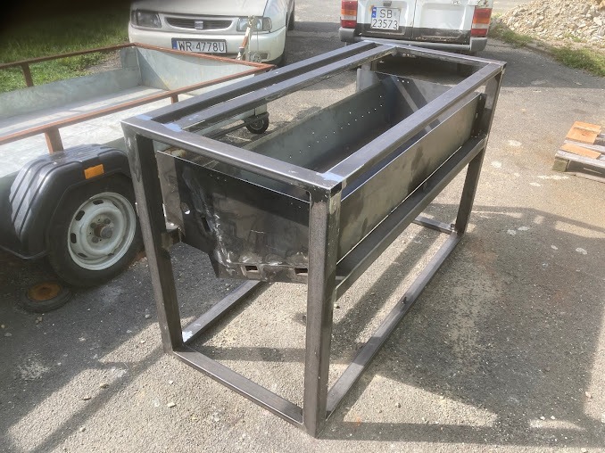 Metalowa konstrukcja w trakcie spawania, widoczne spawy na metalowych profilach, w tle przyczepa i dwa samochody.