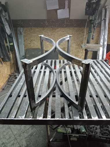 Dwie metalowe, surowe nogi do stołu o łukowatym kształcie, ustawione na metalowym stole warsztatowym z widocznymi śladami obróbki.