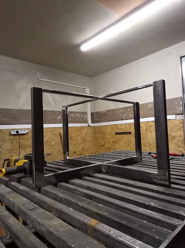 Surowa, metalowa konstrukcja w kształcie sześcianu, spawana na metalowym stole warsztatowym w jasnym oświetleniu, szlifierka kątowa leży obok.