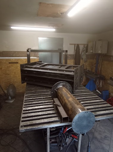 Warsztatowe ujęcie metalowej konstrukcji, rury z flanszą i profili na stole spawalniczym z widocznym wentylatorem i oświetleniem jarzeniowym.