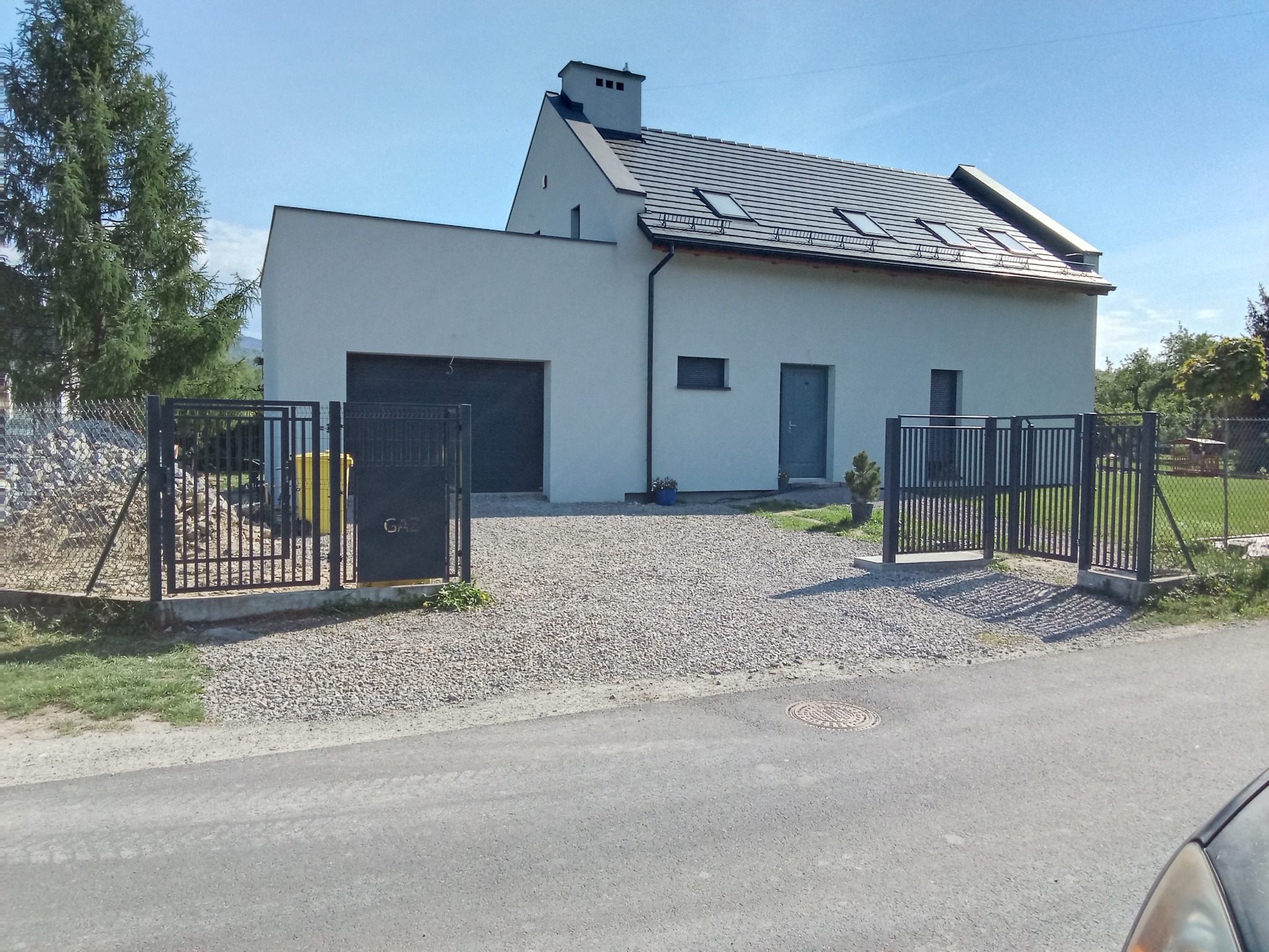 Nowoczesny dom jednorodzinny z szarą bramą wjazdową i furtką. Na furtce widoczny napis 'GAZ'. Podjazd wyłożony drobnym kamieniem.