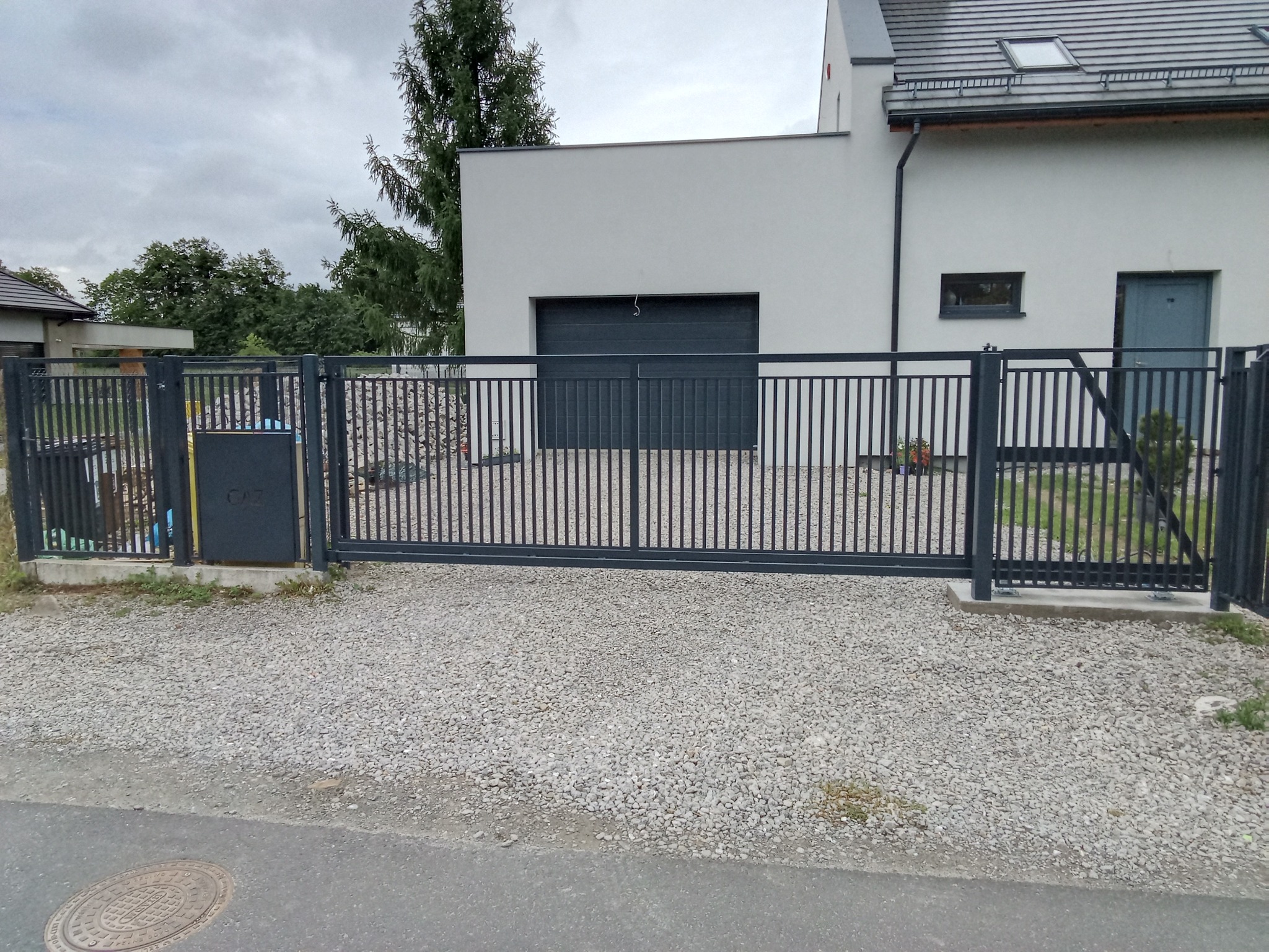 Nowoczesna, przesuwna brama wjazdowa w kolorze grafitowym, z pionowymi szczeblami, na tle domu z garażem i szarą nawierzchnią z kamienia.
