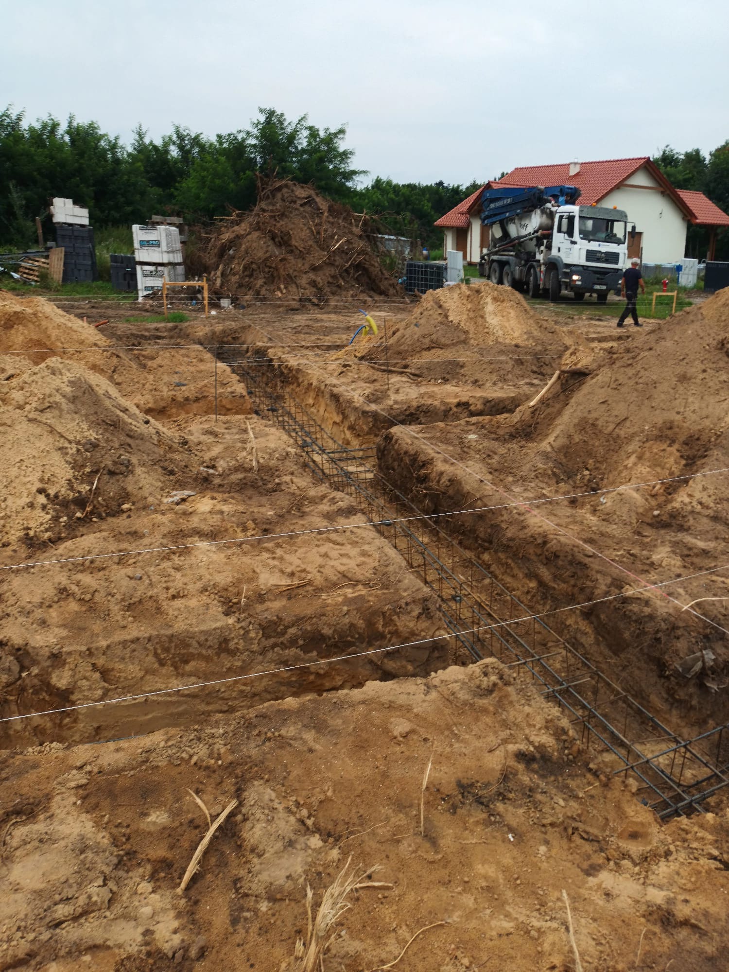 Przygotowany wykop pod fundamenty z widocznym zbrojeniem, na tle domu i betoniarki. Ujęcie z ziemi.