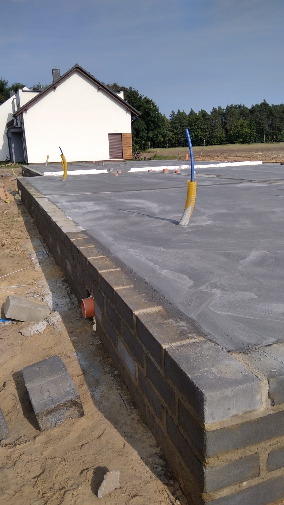 Wylany fundament z wyprowadzonymi instalacjami w Gronówku. Widoczny mur oporowy z pustaków, podłoże piaskowe i fragment domu w tle. Ujęcie z dołu.