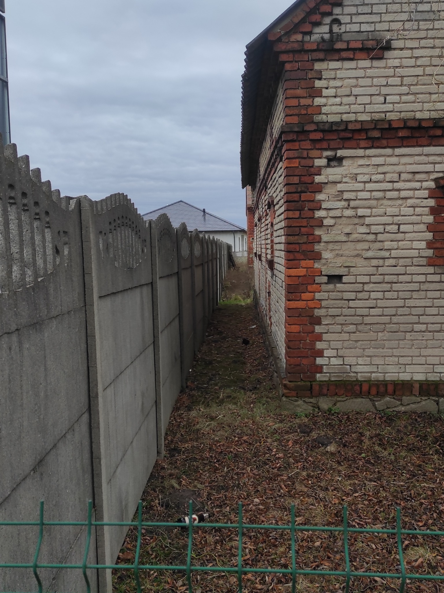 Granica działki z betonowym ogrodzeniem i ceglanym budynkiem w tle, liście na ziemi. Widok na wąską ścieżkę i dom z szarym dachem.