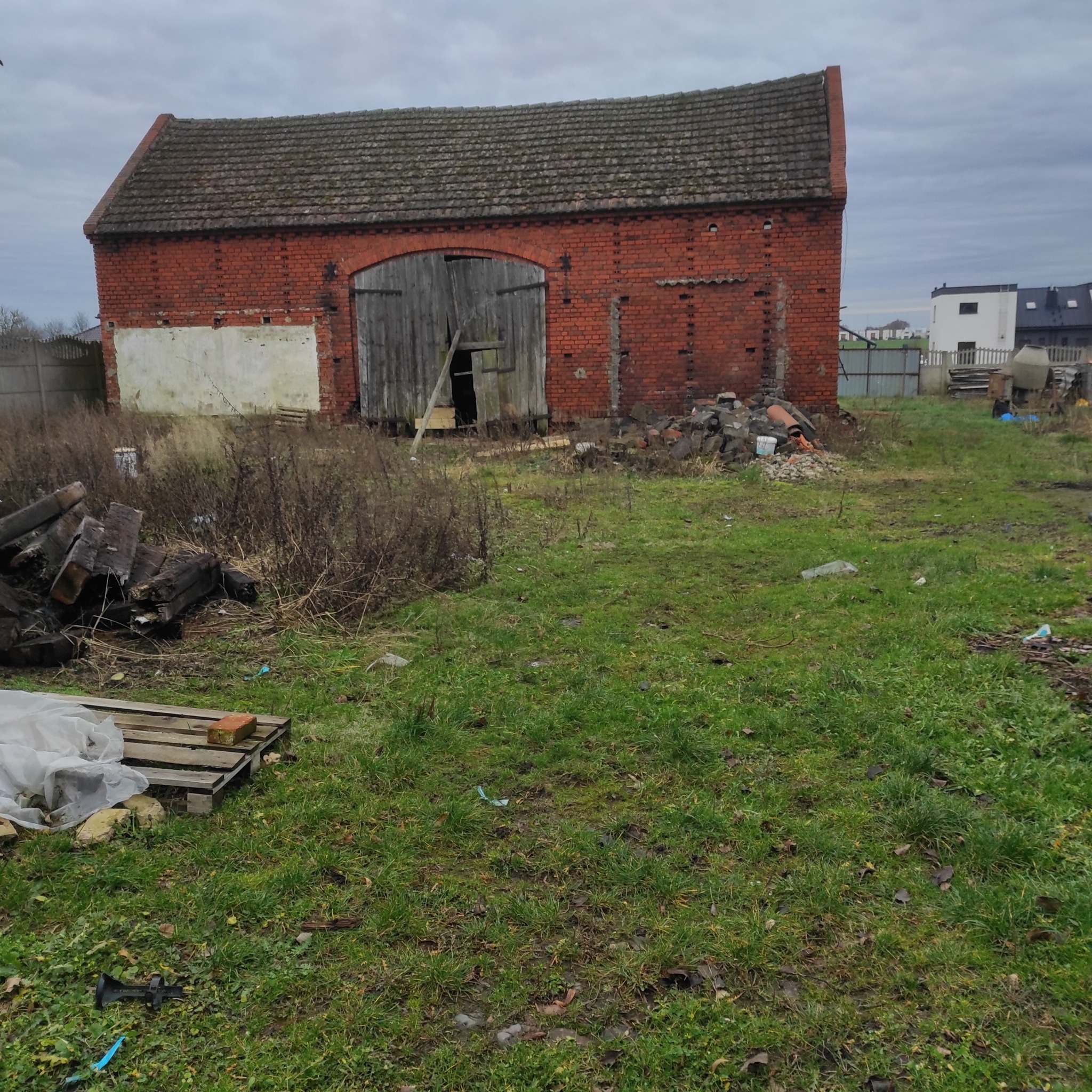 Ceglany budynek gospodarczy z drewnianymi wrotami, otoczony zielenią i gruzem. Widoczne ślady częściowej rozbiórki i zaniedbania. Pochmurne niebo w tle.