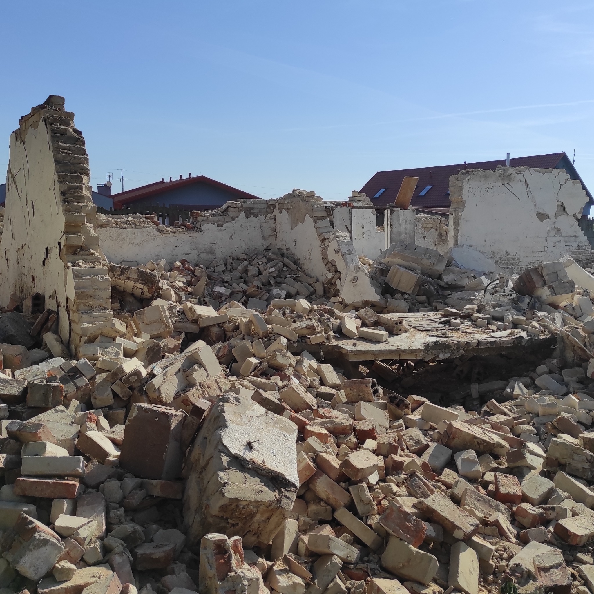 Gruzowisko po rozbiórce budynku z fragmentami ścian, cegłami i elementami konstrukcyjnymi na tle błękitnego nieba i zabudowań mieszkalnych.