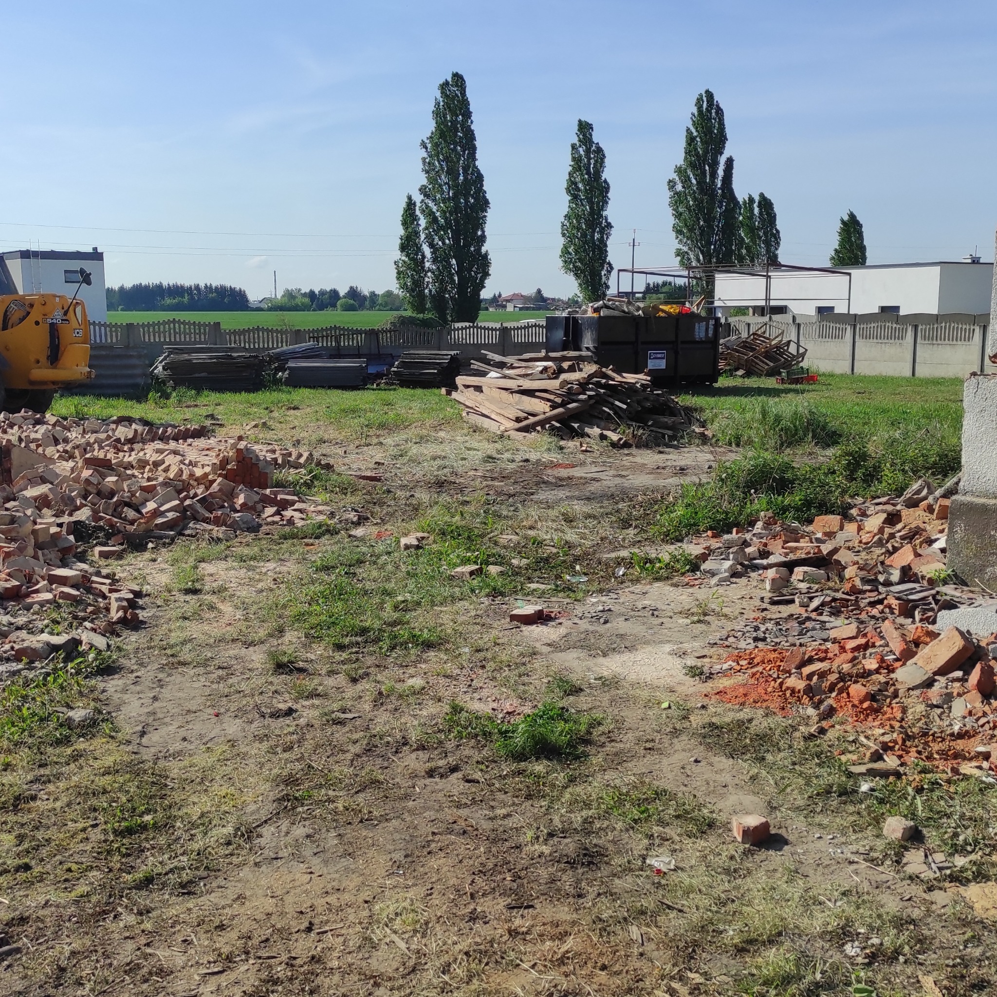 Teren po rozbiórce: gruz ceglany, deski, kontener na odpady budowlane. W tle widoczne drzewa i zabudowania.