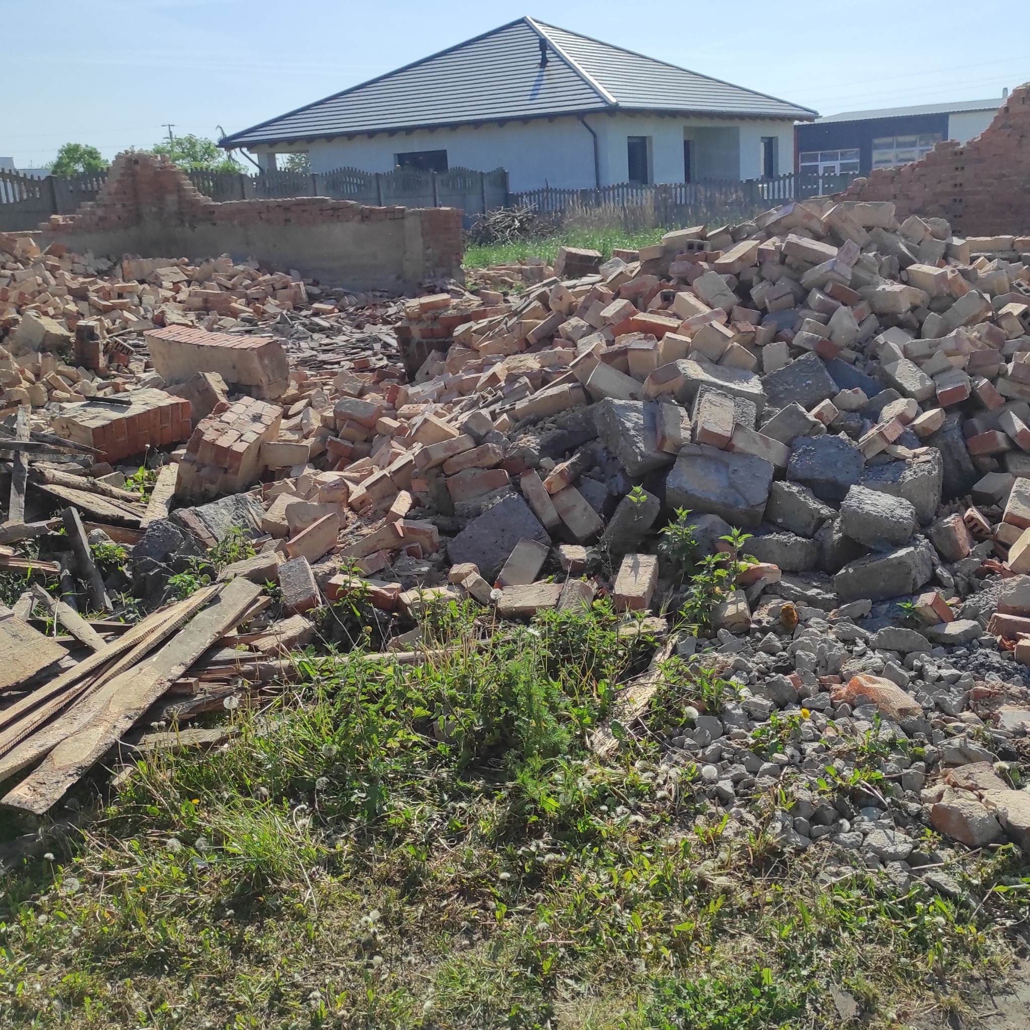 Gruz ceglany i betonowy po rozbiórce, z zielenią i elementami drewnianymi na pierwszym planie, w tle budynek z szarą dachówką i fragment zrujnowanej ściany.