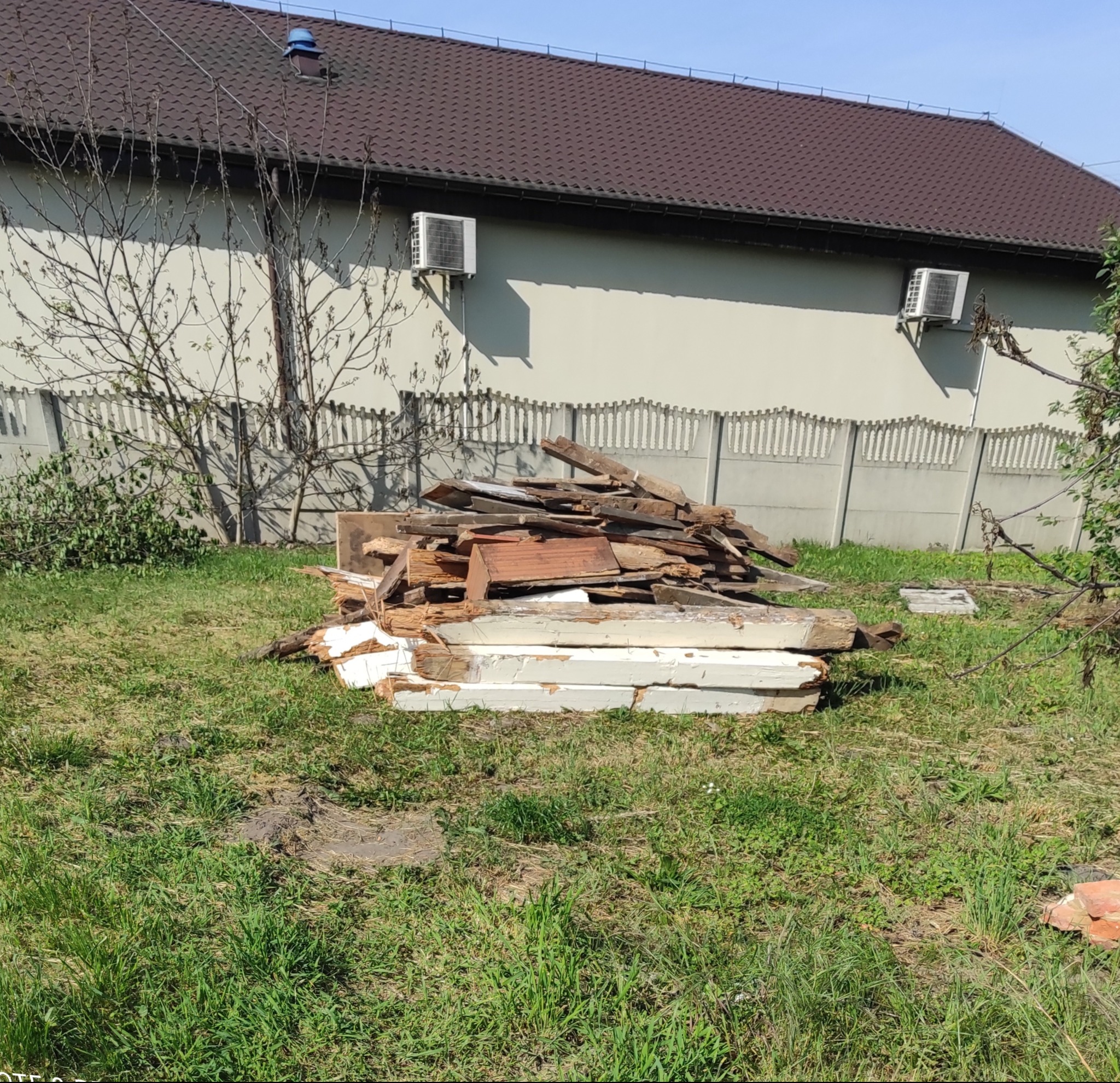 Stos drewnianych odpadów budowlanych na trawie przed domem z szarą elewacją i brązowym dachem. Widoczne klimatyzatory na ścianie i betonowe ogrodzenie.