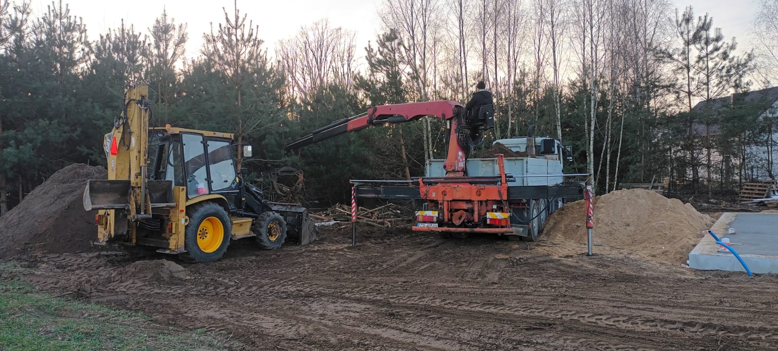 Żółta koparko-ładowarka i ciężarówka z dźwigiem podczas prac ziemnych na działce z fundamentami i stertami ziemi. W tle drzewa.