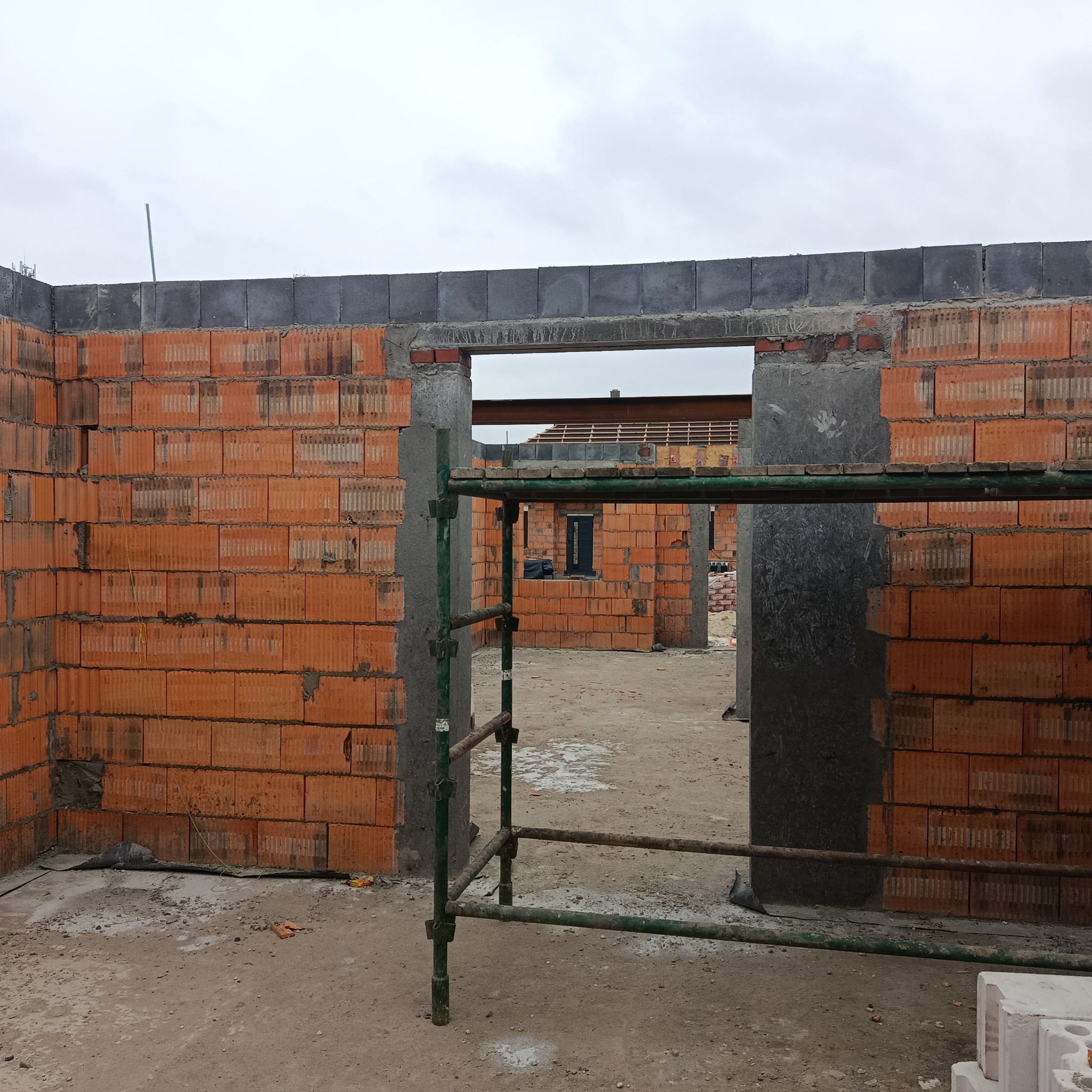 Wnętrze domu w trakcie budowy: ściany z czerwonej cegły, widoczne rusztowanie i otwory okienne. W tle konstrukcja dachu i materiały budowlane.