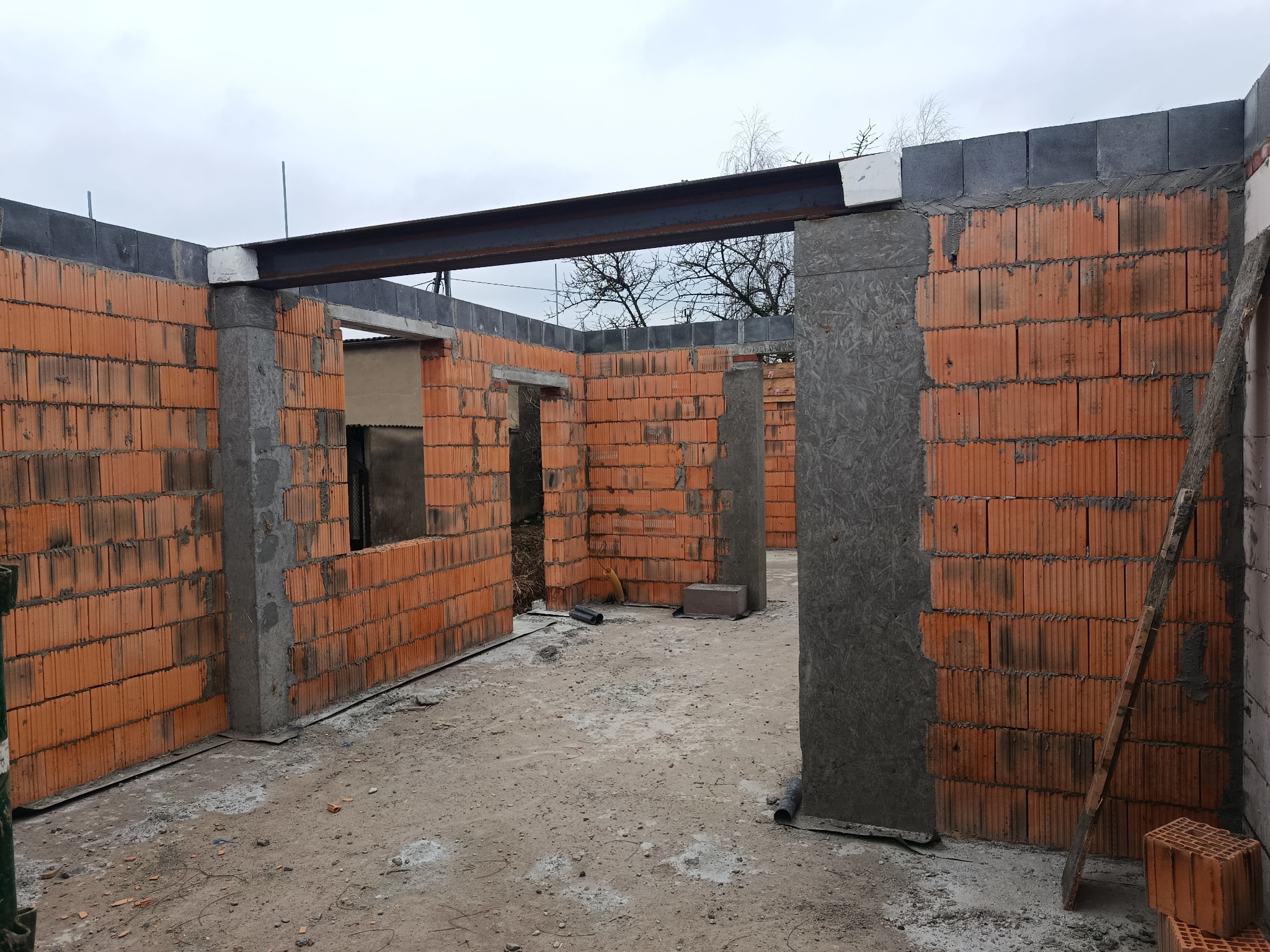 Surowy stan domu w trakcie budowy: ściany z czerwonej cegły, betonowe słupy, stalowa belka stropowa. Widoczne otwory okienne i drzwiowe, szary betonowy fundament.