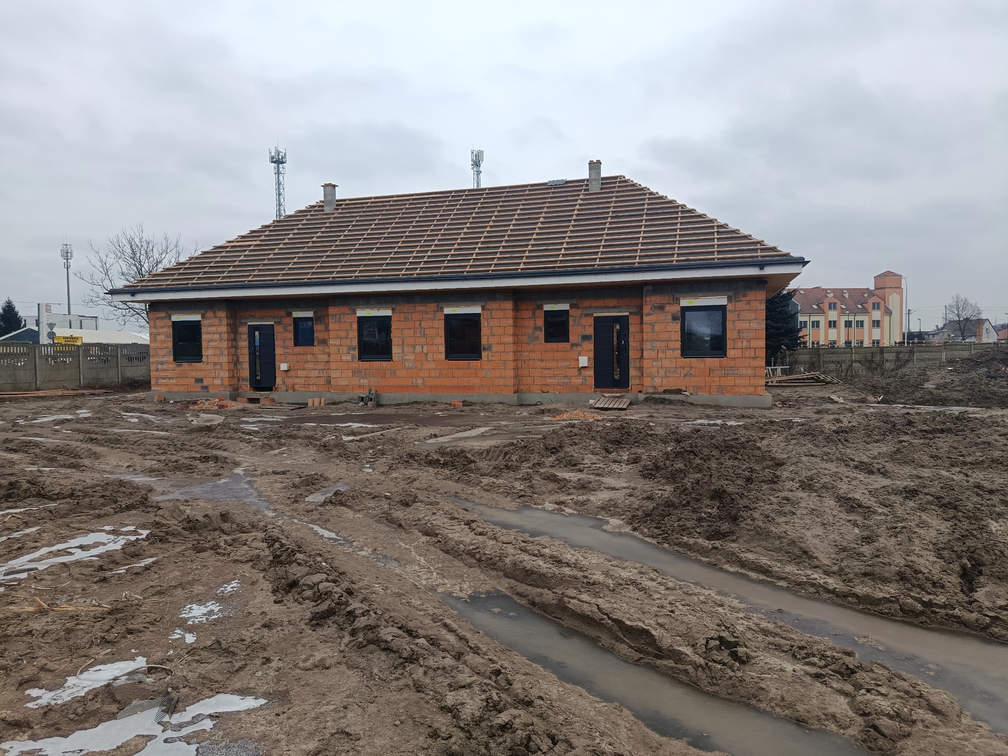 Surowy, ceglany dom w trakcie budowy z częściowo ułożonym dachem z dachówki, czarnymi oknami i drzwiami. Teren wokół rozjeżdżony, błotnisty. Pochmurne niebo w tle.