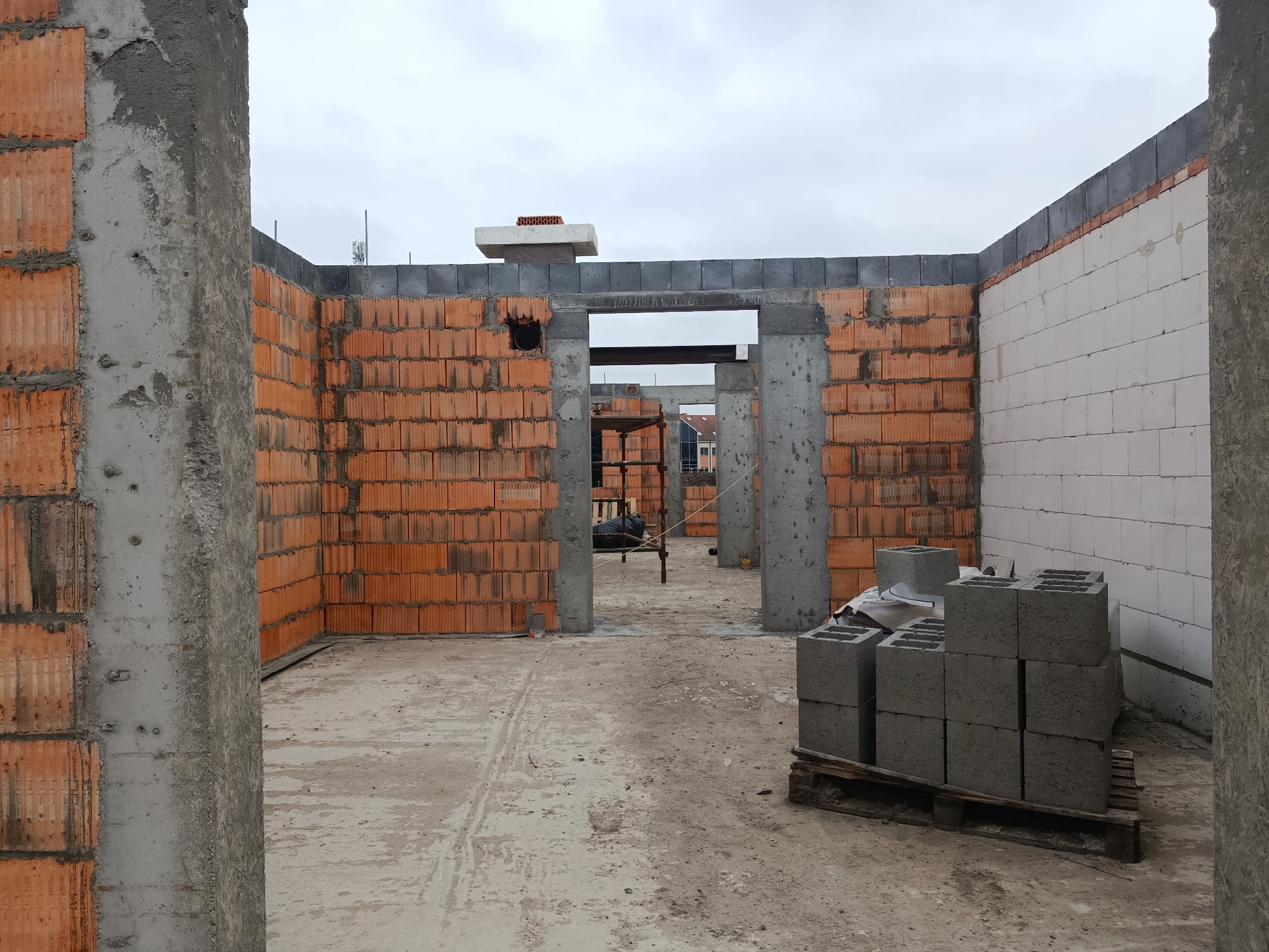 Wnętrze domu w trakcie budowy: ściany z czerwonej cegły i białych bloczków, betonowe filary, paleta z szarymi bloczkami. Widoczne rusztowania i elementy konstrukcyjne.