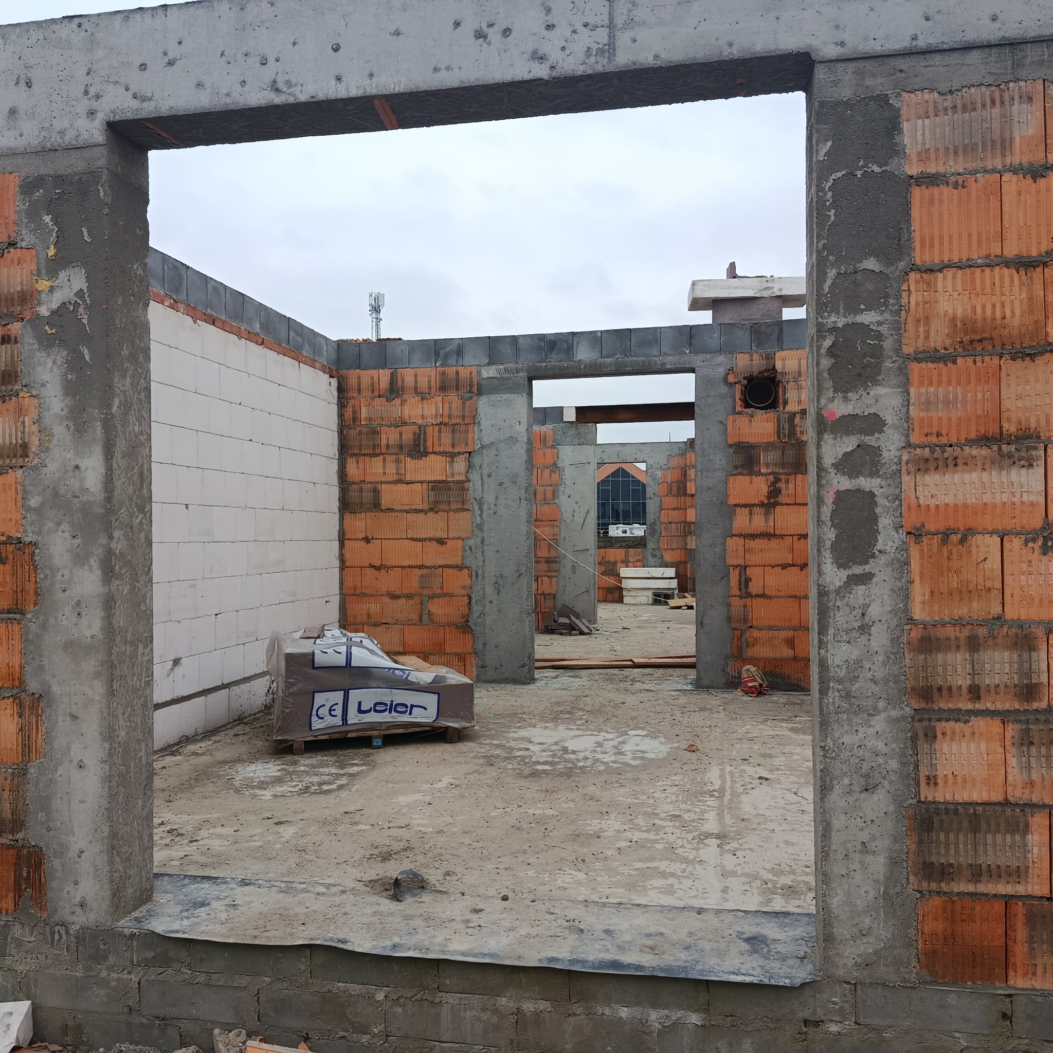 Surowy stan domu murowanego w trakcie budowy. Widoczne ściany z czerwonej cegły i bloczków, betonowe nadproża i słupy. W tle budynek z przeszkloną fasadą.