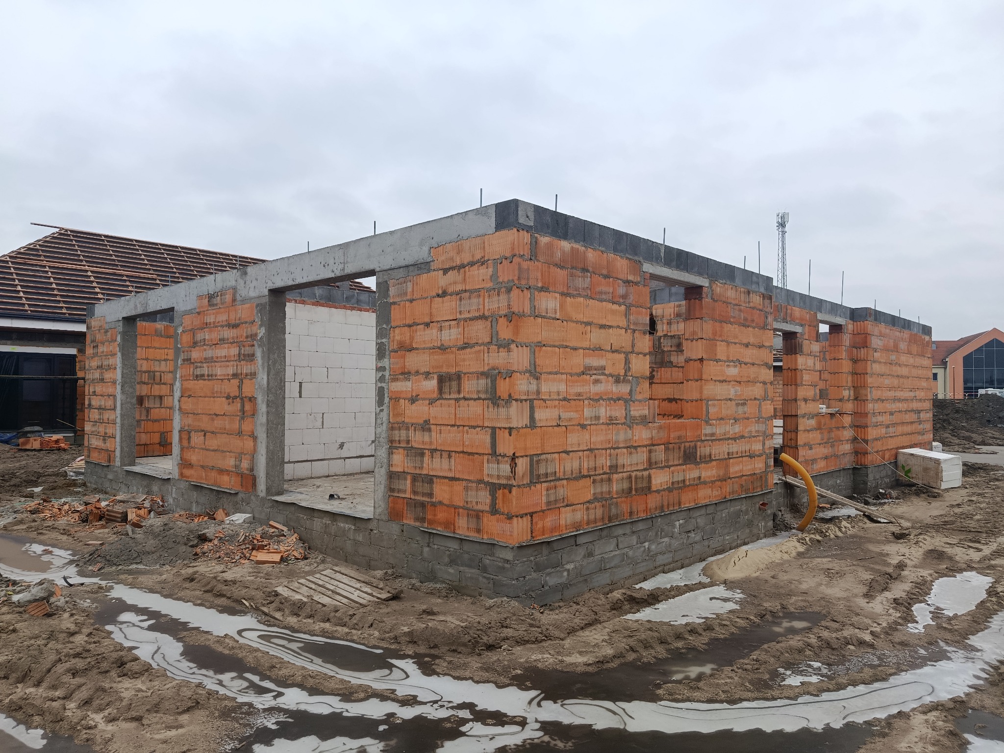 Surowy stan domu z czerwonej cegły i betonu pod szarym niebem. Widoczne fundamenty i ściany w trakcie budowy. W tle zarys dachu sąsiedniego budynku.