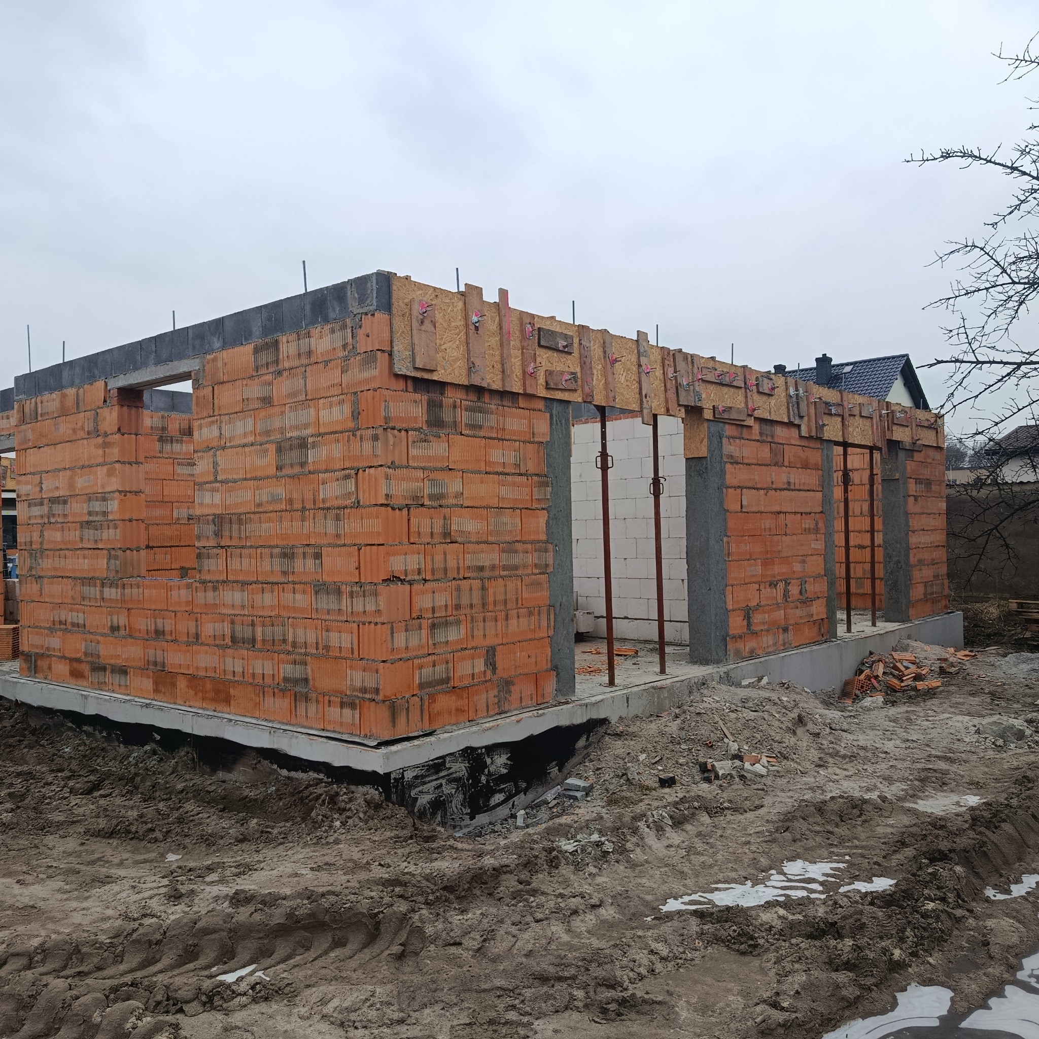 Surowy stan domu z czerwonej cegły, widoczne szalunki i podpory, fundamenty zabezpieczone czarną izolacją, ziemia wokół, pochmurne niebo w tle.