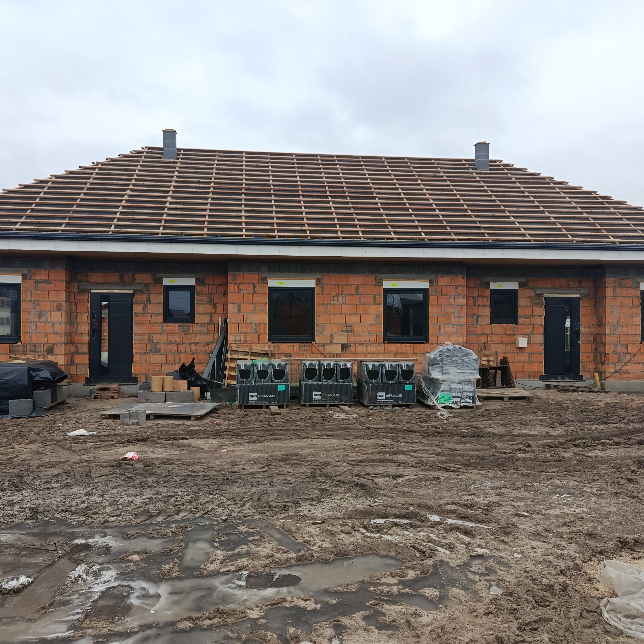 Budowa domu z czerwonej cegły w trakcie realizacji. Widoczne czarne okna i drzwi, drewniana konstrukcja dachu oraz materiały budowlane na błotnistym terenie.