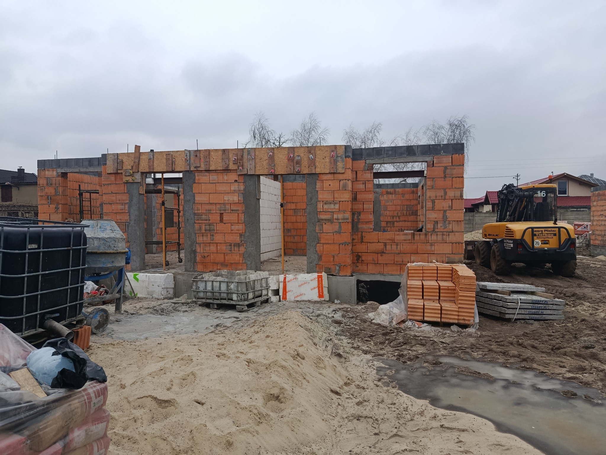 Wznoszenie ścian parteru domu z czerwonej cegły, widoczny żółty podnośnik teleskopowy Mecalac i materiały budowlane na placu budowy w pochmurny dzień.