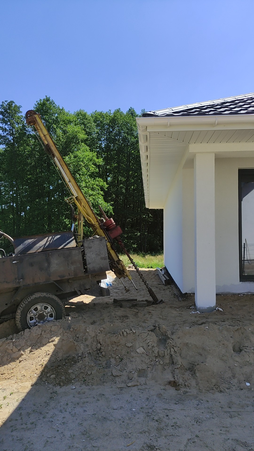 Żółta wiertnica do gruntu obok białego domu z czarnym dachem, widoczny wykop i drzewa w tle.