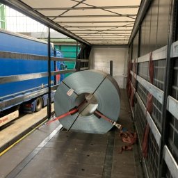 Zaratustra Sp. z o. o. - Wnętrze naczepy z zabezpieczoną rolą blachy stalowej, widoczne pasy mocujące i fragment niebieskiej ciężarówki załadunkowej. Bezpieczny transport materiałów.