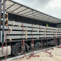 Zaratustra Sp. z o. o. - Ciężarówka z otwartą naczepą, zabezpieczona ładunkiem stalowych elementów na paletach, spiętych pasami transportowymi. Widoczny pracownik w kamizelce odblaskowej.