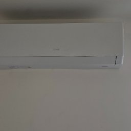 Leopold Molski Notos Klima - Biały klimatyzator marki Gree zamontowany na jasnej ścianie w pomieszczeniu, obok okna zabezpieczonego folią malarską.