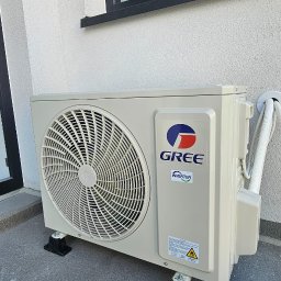 Leopold Molski Notos Klima - Zewnętrzna jednostka klimatyzacji Gree zamontowana na czarnych podstawkach, widoczne rury i okno z folią ochronną.