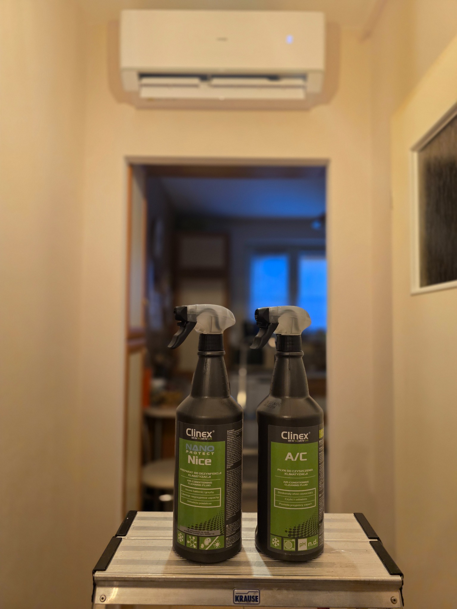 Dwa spraye Clinex A/C do czyszczenia klimatyzacji, ustawione na aluminiowej drabinie, z zamontowaną klimatyzacją na ścianie w tle.