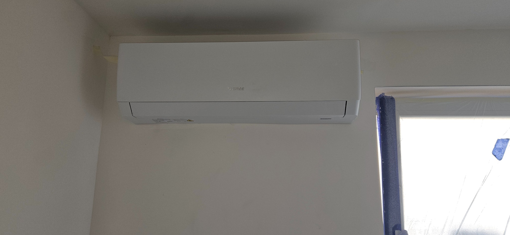 Biały klimatyzator marki Gree zamontowany na jasnej ścianie w pomieszczeniu, obok okna zabezpieczonego folią malarską.