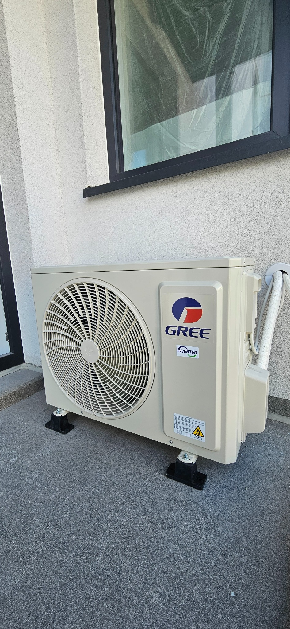 Zewnętrzna jednostka klimatyzacji Gree zamontowana na czarnych podstawkach, widoczne rury i okno z folią ochronną.