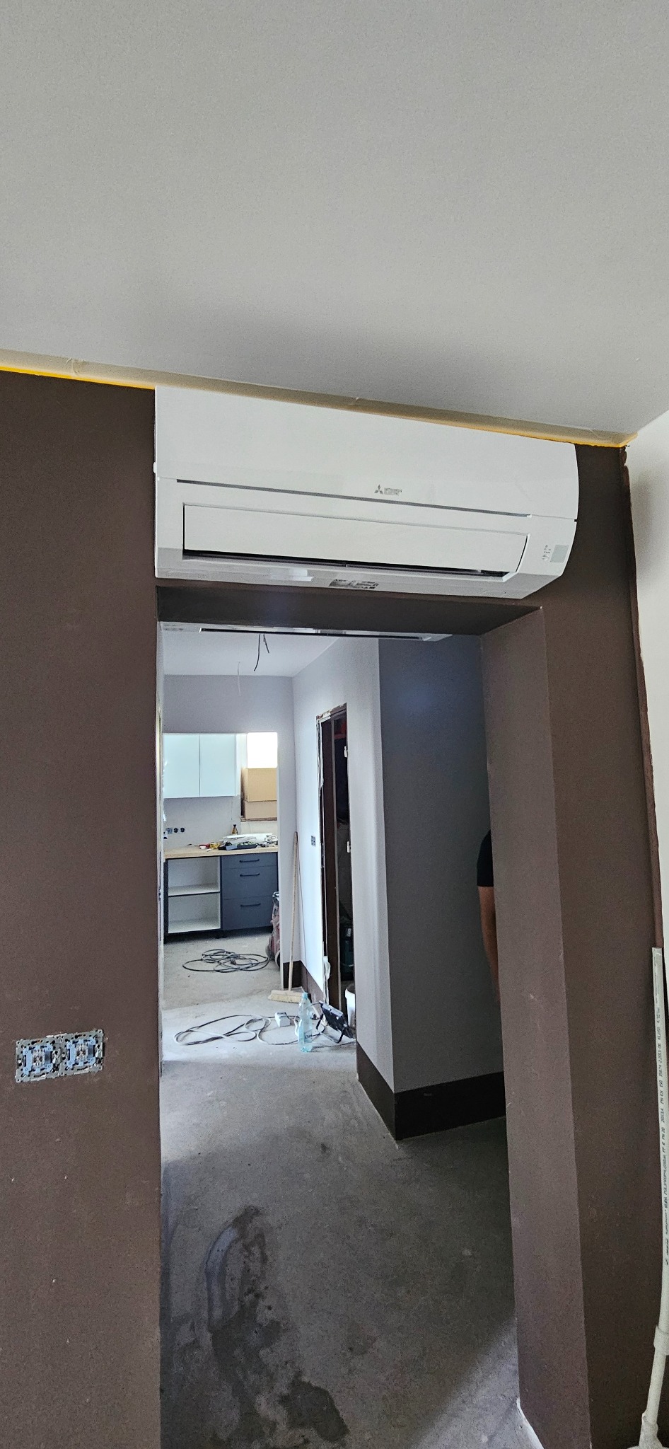 Biała jednostka klimatyzacyjna Mitsubishi zamontowana nad wejściem do pokoju z widoczną kuchnią w tle podczas remontu.