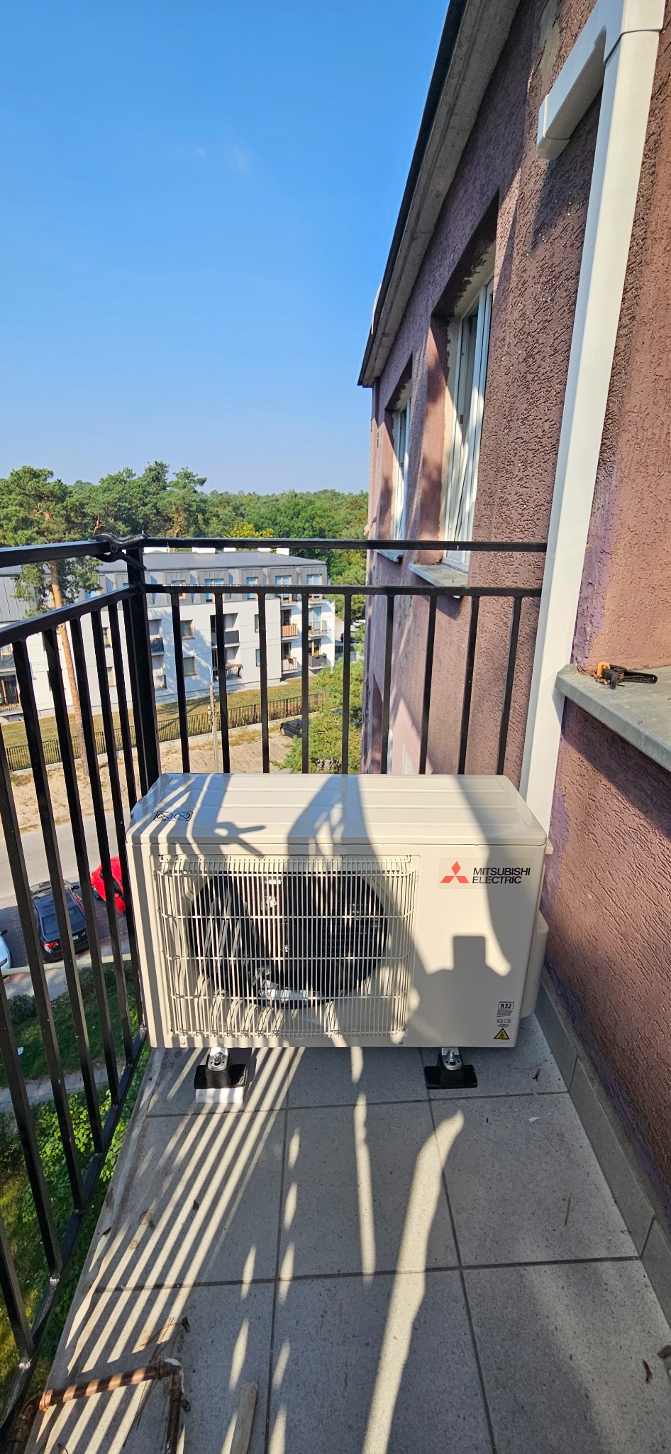 Jednostka zewnętrzna klimatyzatora Mitsubishi Electric zamontowana na balkonie w bloku mieszkalnym, widok z góry, cień rzucany przez balustradę.