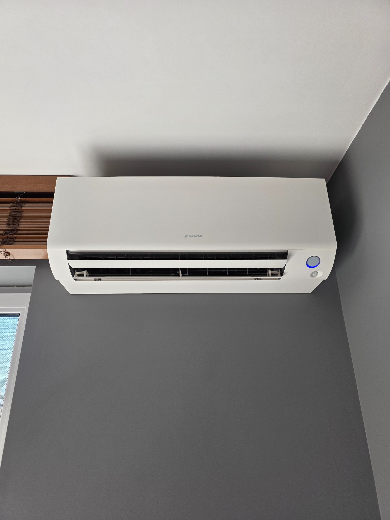 Biała jednostka wewnętrzna klimatyzacji Daikin zamontowana na szarej ścianie w pomieszczeniu, widoczne żaluzje okienne w tle.