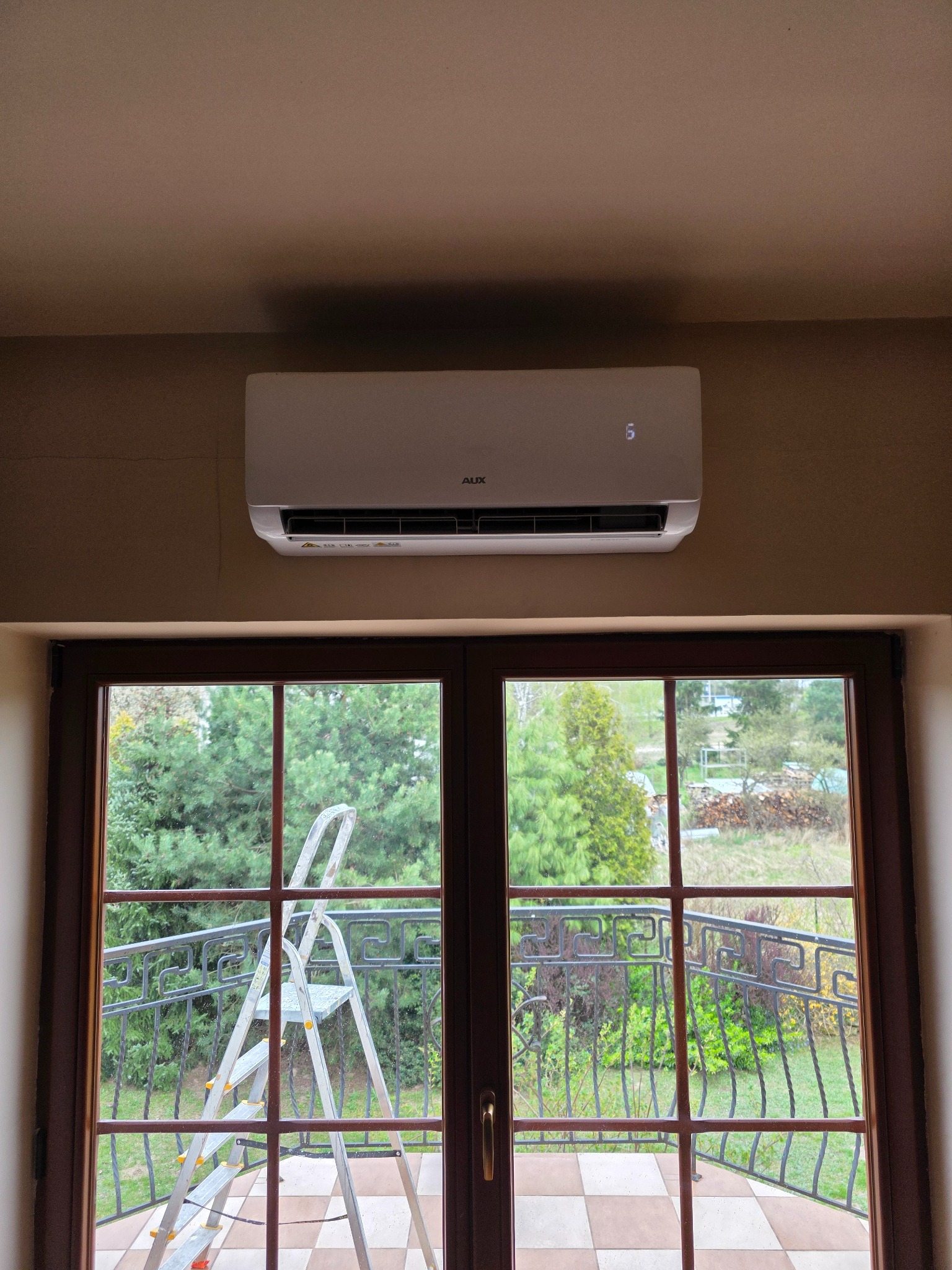 Biały klimatyzator AUX zamontowany nad dwuskrzydłowymi drzwiami balkonowymi, z widoczną drabiną opartą o balustradę balkonu i zieleń w tle.