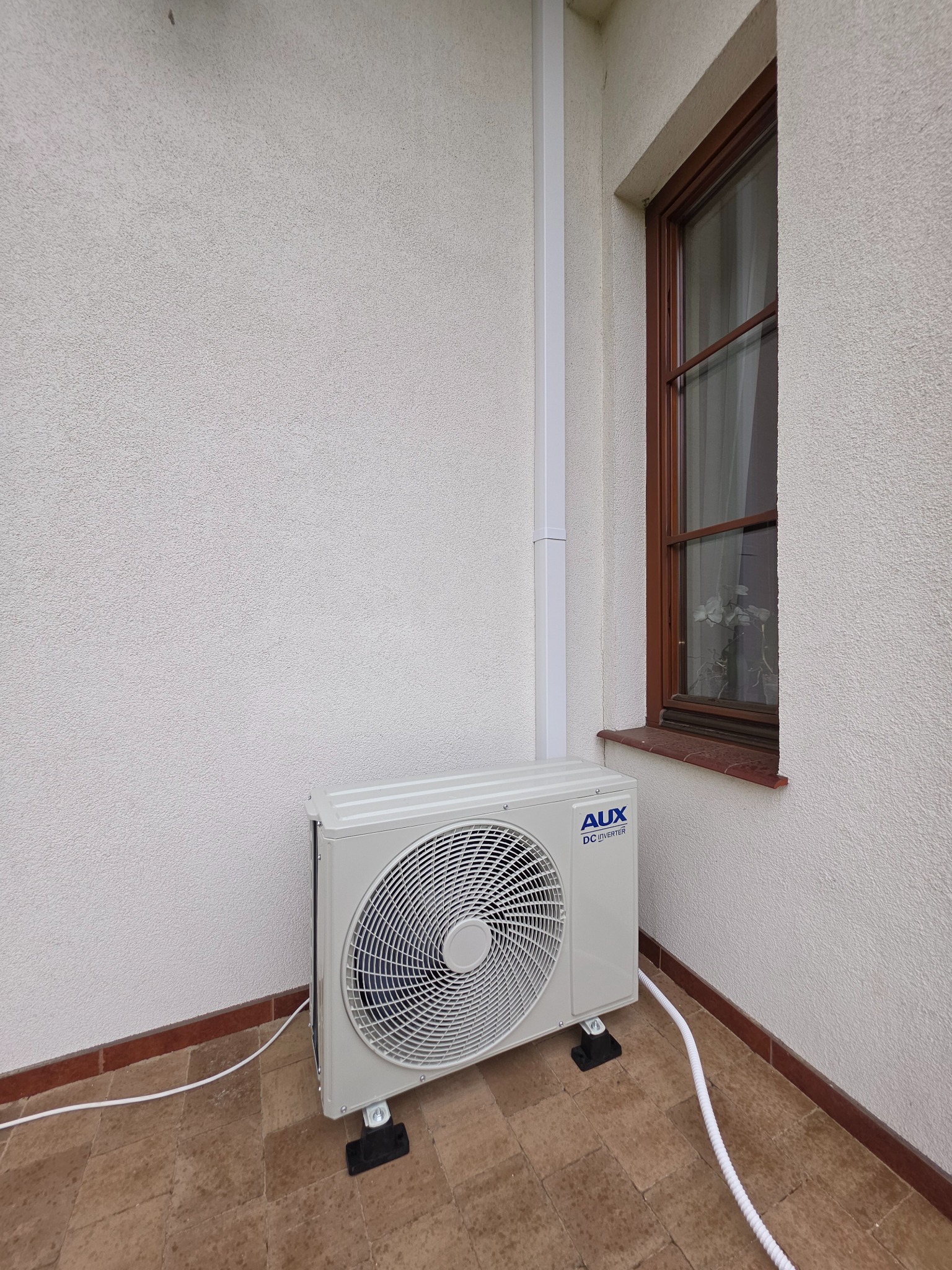 Zewnętrzna jednostka klimatyzacji AUX DC Inverter umieszczona na tarasie obok okna z brązową ramą i białej ściany z rurą spustową.