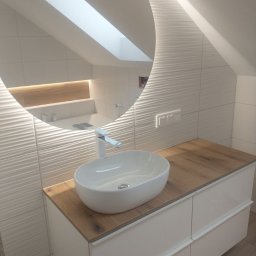 PK-Progres - Biała, owalna umywalka nablatowa z chromowaną baterią na blacie z drewnopodobnego laminatu, podwieszona szafka z białymi, lakierowanymi frontami, ściana z białych, falistych płytek, okrągłe lustro...