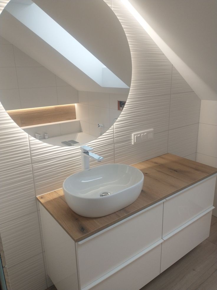 Biała, owalna umywalka nablatowa z chromowaną baterią na blacie z drewnopodobnego laminatu, podwieszona szafka z białymi, lakierowanymi frontami, ściana z białych, falistych płytek, okrągłe lustro...