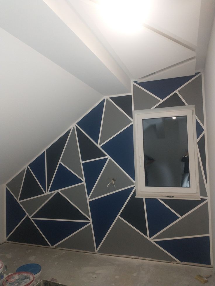Ściana w pokoju na poddaszu pomalowana w geometryczne wzory z użyciem farb w odcieniach szarości i niebieskiego, oddzielone białymi liniami, widoczne puszki z farbą na podłodze.